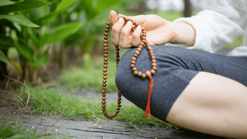 Pessoa meditando sentada na grama segurando um colar de contas