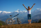 Mulher em topo de montanha, ao lado de uma bicicleta. Ela está de pé e com os braços esticados para o alto.