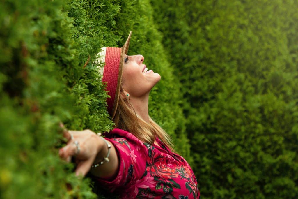 Mulher encostada em uma parede de plantas, sorrindo.