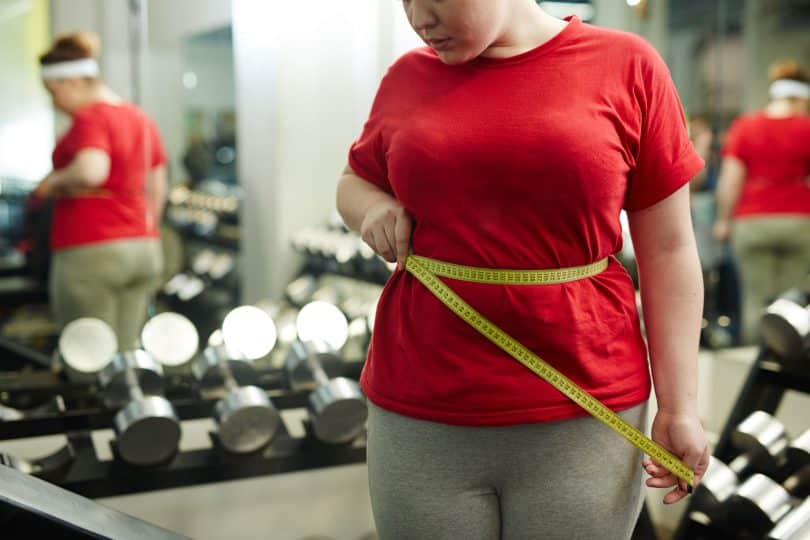 Mulher mede a cintura com fita métrica. O ambiente é de academia.