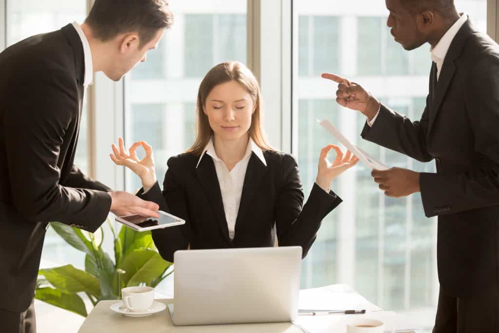 Mulher calma, meditando, enquanto dois homens gritam com ela em ambiente de trabalho