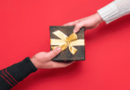 Mão estendendo uma caixa de presente preta com laço dourado para outra pessoa, em fundo vermelho.