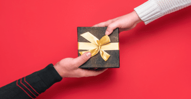 Mão estendendo uma caixa de presente preta com laço dourado para outra pessoa, em fundo vermelho.