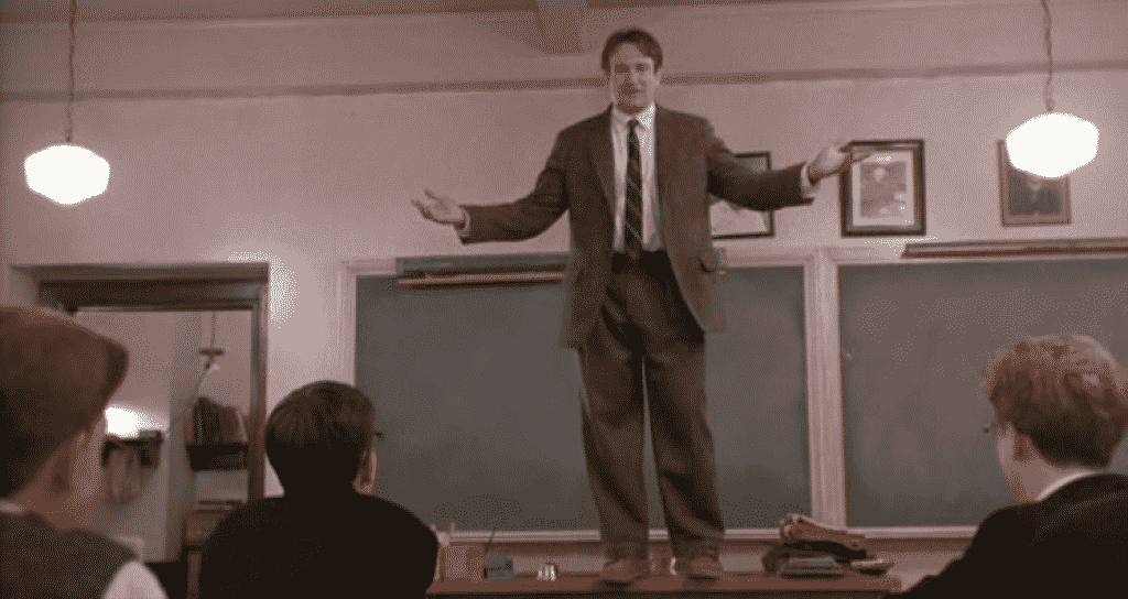 Professor protagonista do filme, John Keating, em pé sobre uma mesa em uma sala de aula, conversando com seus alunos.