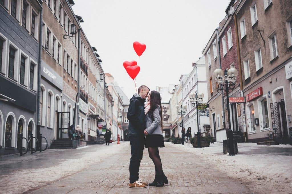 Casal segurando balões em formato de coração enquanto se beijam no meio da rua.