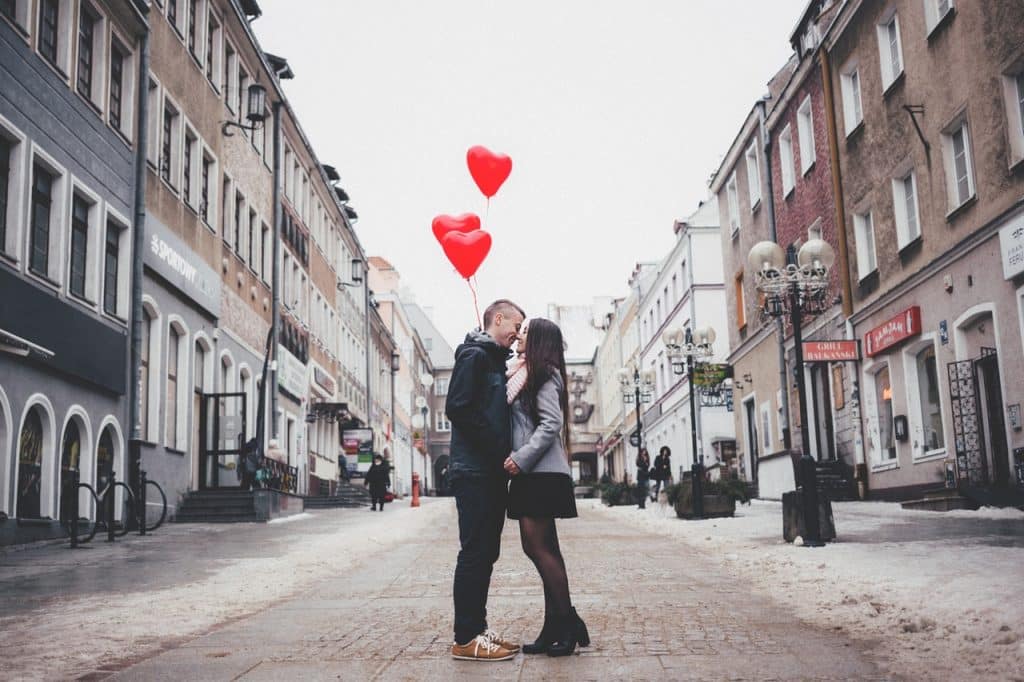 Casal segurando balões em formato de coração enquanto se beijam no meio da rua. 