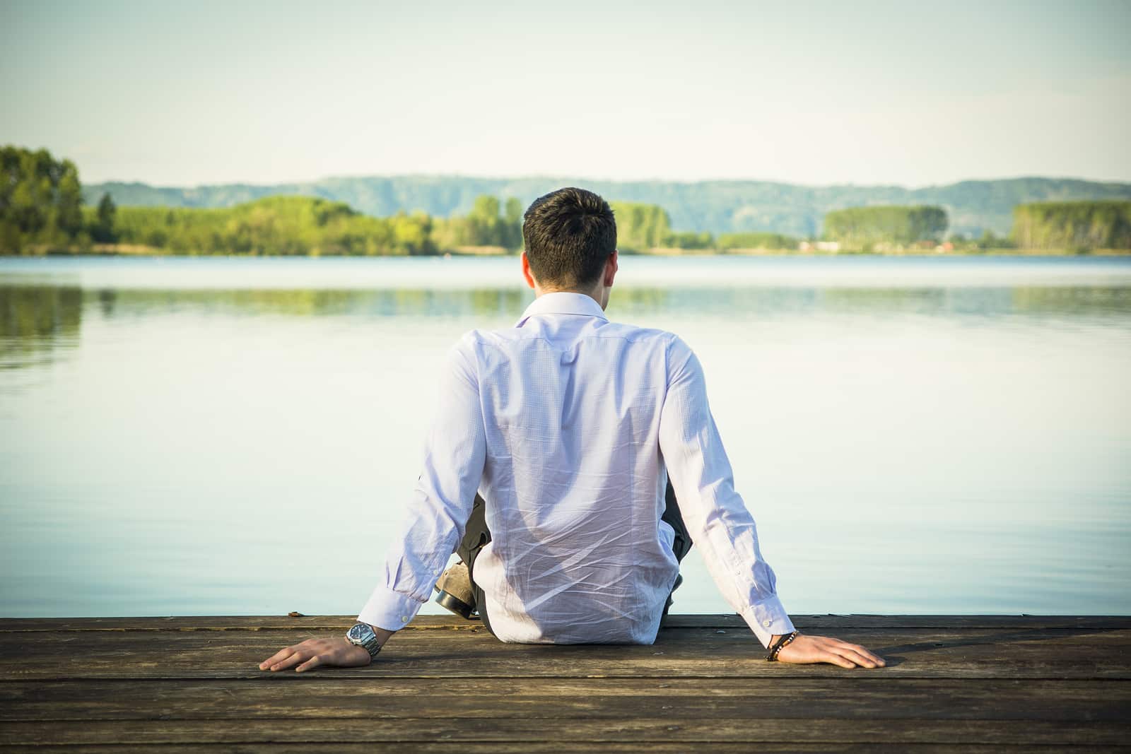 Handsome young man on a lake in a sunny, peaceful day, sitting on a wood pier, thinking or meditating