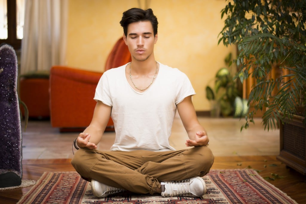 Young man meditating on his living room floor, sitting in the lotus position with his eyes closed and an expression of tranquility, in a health and fitness concept