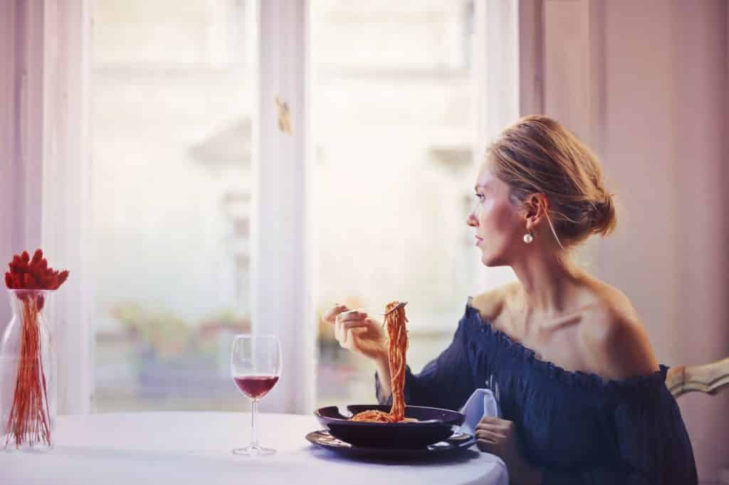 Mulher comendo macarrão e bebendo uma taça de vinho enquanto olha pela janela.