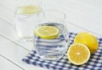 Dois copos com água e uma rodela de limão siciliano em cada. Um dos copos está sobre um pano xadrez. Ao lado, um limão siciliano inteiro, e metade de outro.