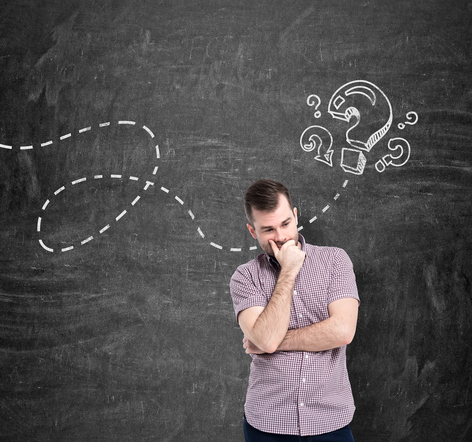 A young man in casual shirt is thinking about unanswered questions. Question marks are drawn on the black chalkboard.