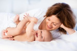 Mother and child on a white bed. Mom and baby boy in diaper playing in sunny bedroom. Parent and little kid relaxing at home. Family having fun together. Bedding and textile for infant nursery.