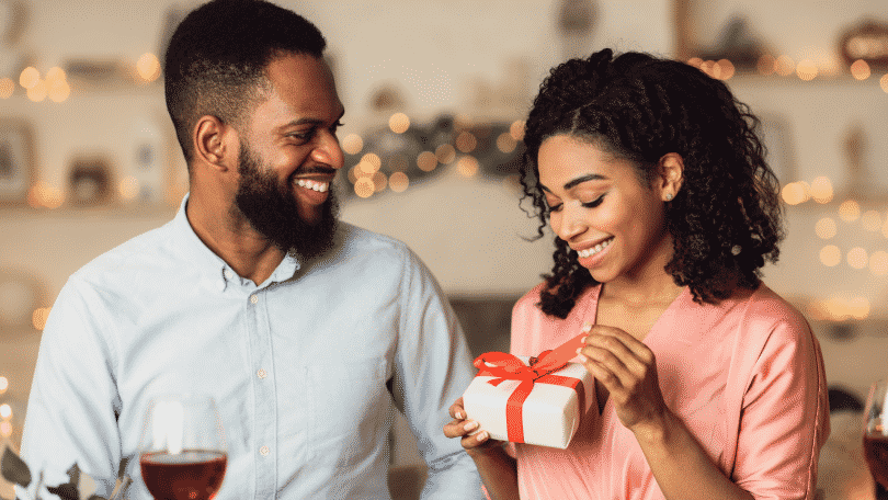 Mulher abrindo presente que o namorado deu. Os dois estão sorrindo e sentados em frente a uma mesa de jantar