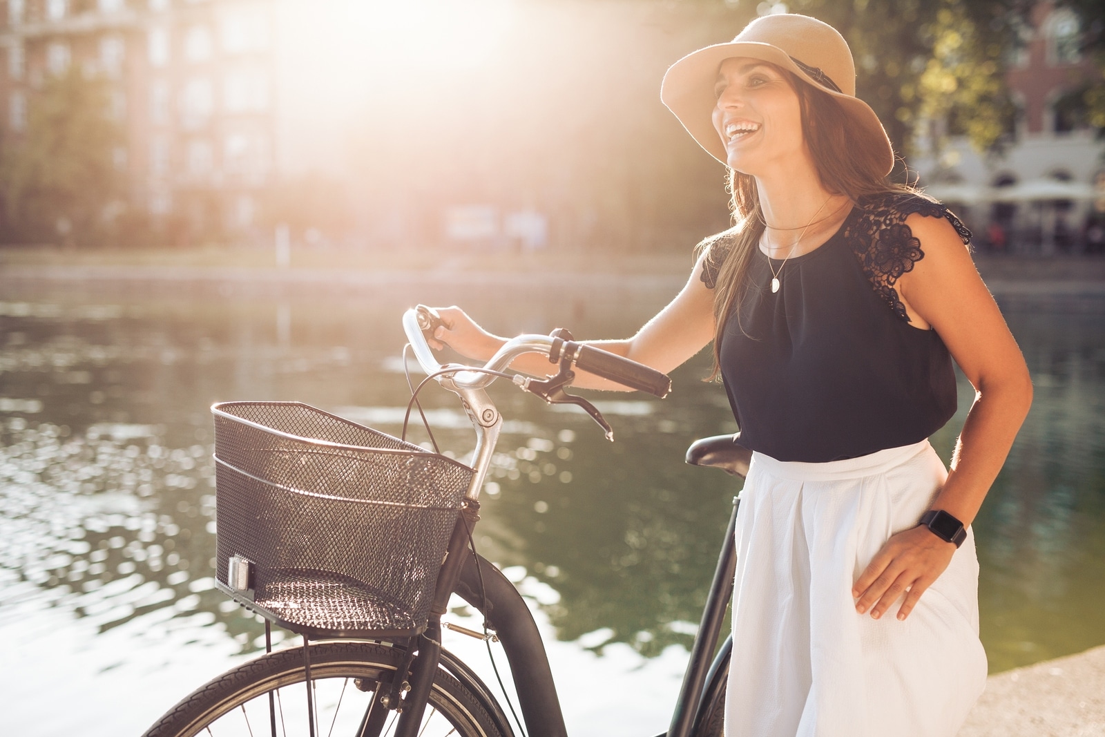 Portrait of beautiful young woman wearing a hat with a bicycle walking along a pond. Happy woman with a bike at the park looking away laughing on a summer day with sun flare.