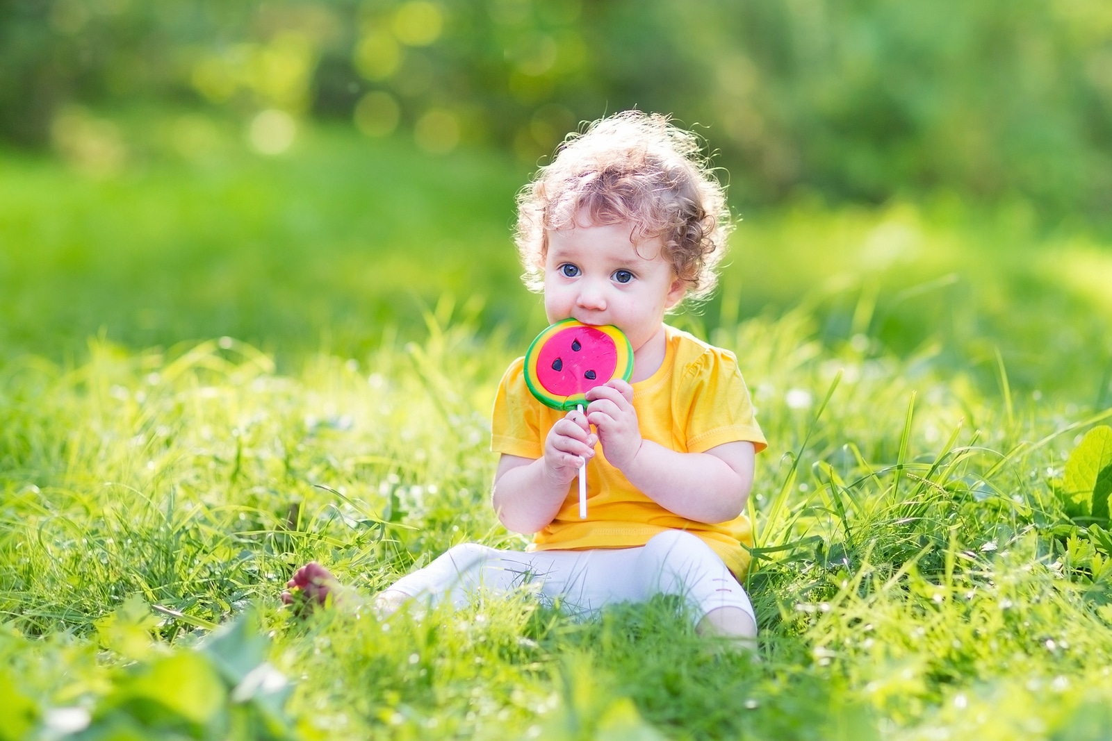 Cute curly baby girl eating watermelon candy in a sunny park
