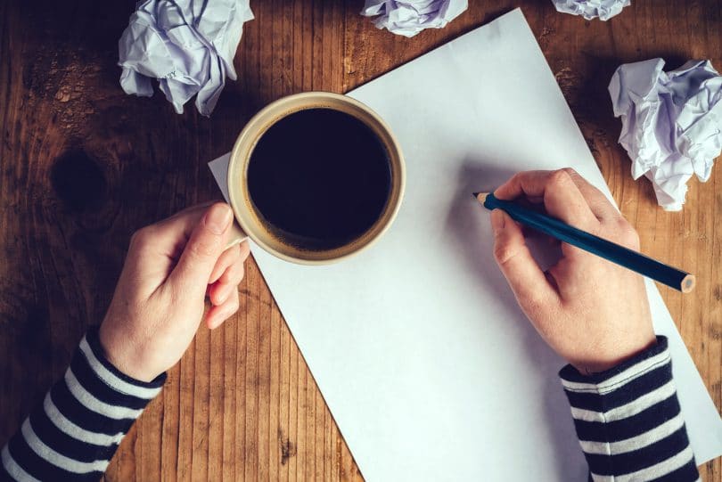 Mãos sobre uma mesa segurando café e escrevendo em um papel sulfite