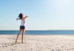 Mulher na praia com braços abertos olhando para o mar