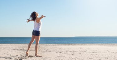 Mulher na praia com braços abertos olhando para o mar