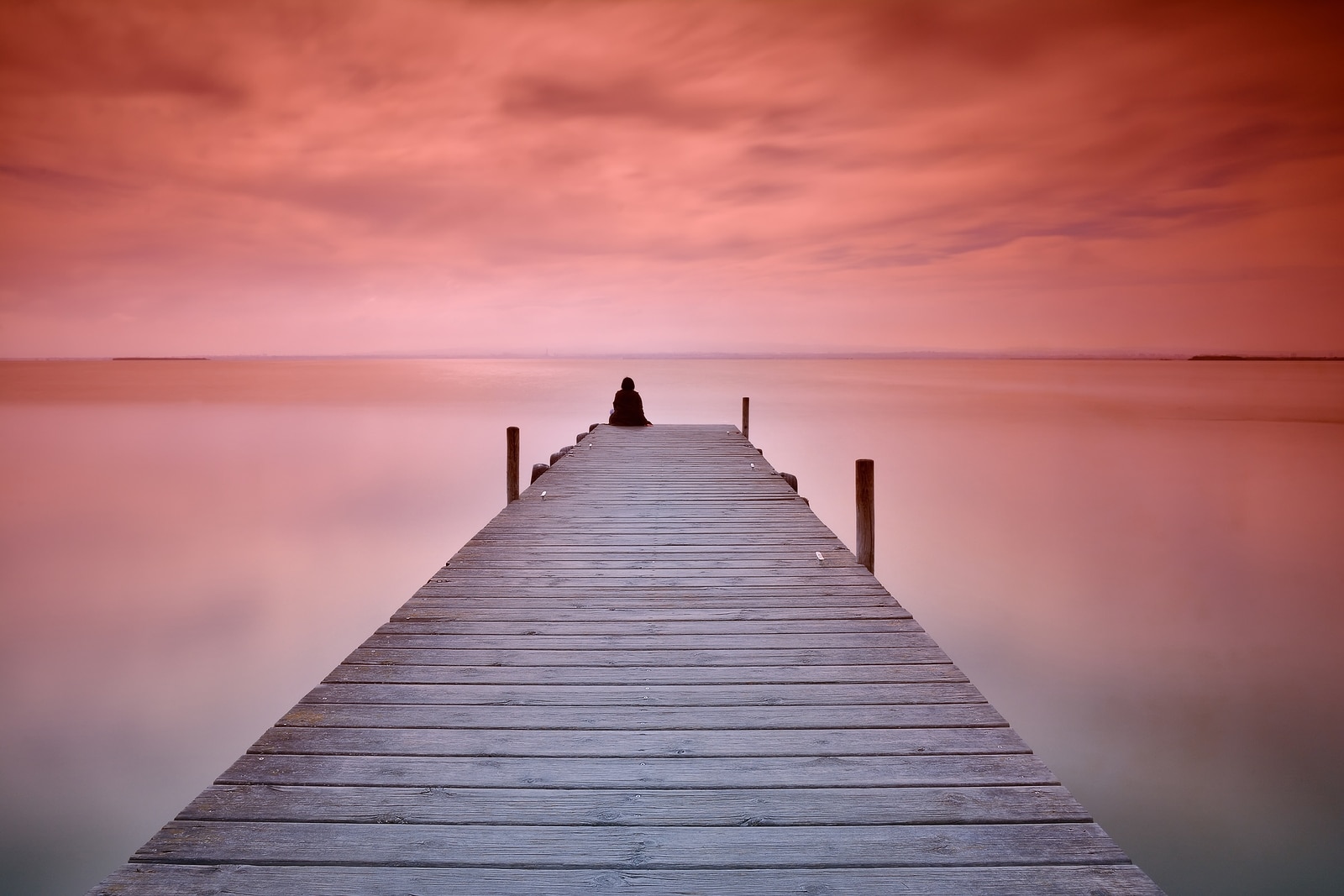 Unrecognizable person sitting on edge of wooden pier at sunset of red and violet colors