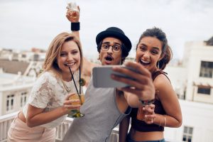 Excited young people taking self portrait with mobile phone during a party. Happy young man and woman taking self portrait at rooftop party. Multiracial people having fun in party with drinks.