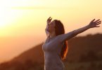 Mulher de braços aberto e sorrindo em campo aberto com a luz do pôr do sol