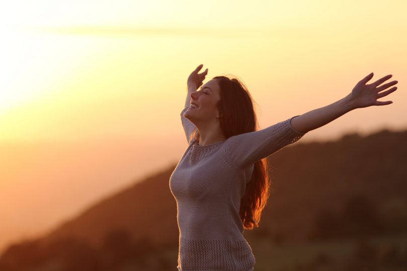 Mulher de braços aberto e sorrindo em campo aberto com a luz do pôr do sol