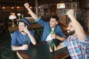 people, leisure, friendship and bachelor party concept - happy male friends drinking bottled beer and raised hands rooting for football match at bar or pub