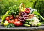 Vegetais e frutas em cesta sobre superfície de madeira escura. O cenário é externo e arborizado.