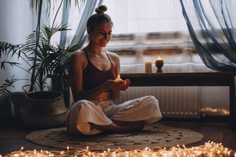 Mulher sorridente se preparando para meditar. Ela segura uma vela acessa e está rodeada por outras velas dispostas no chão em que está sentada.