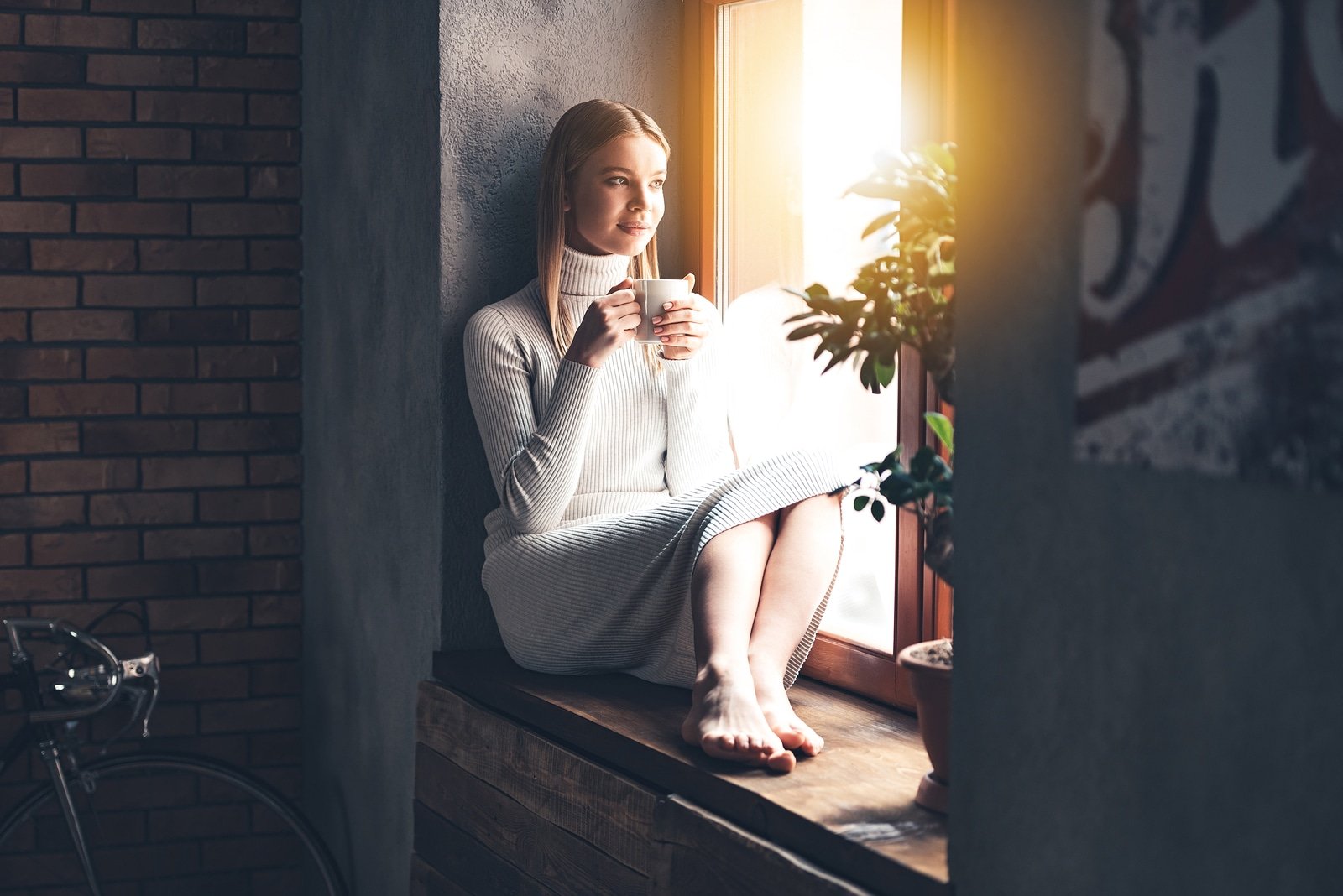 Making plans for new day. Beautiful young woman holding coffee cup and looking through window while sitting at windowsill at home