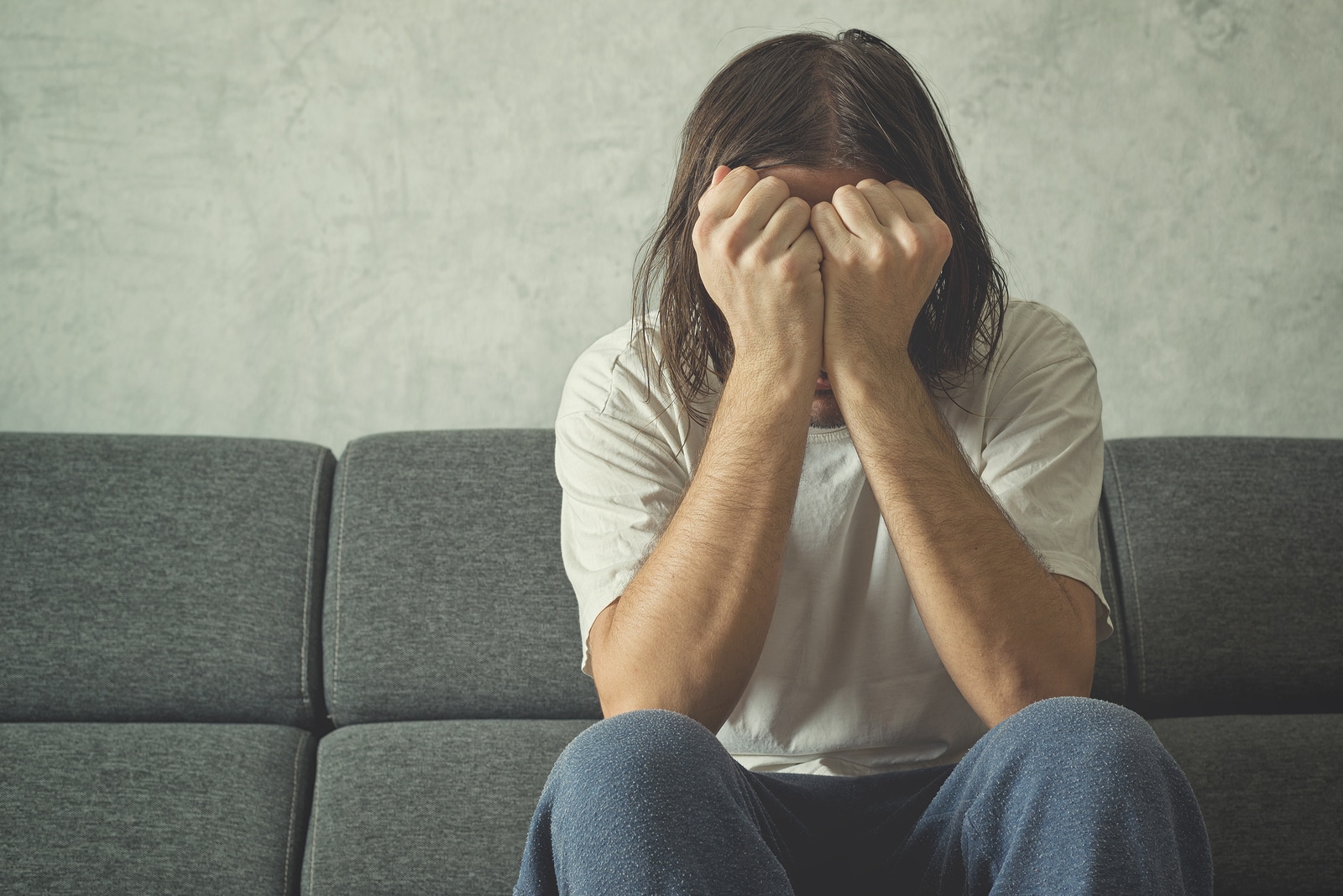 Depressed and sad man on the couch in the room covering face and crying in despair.