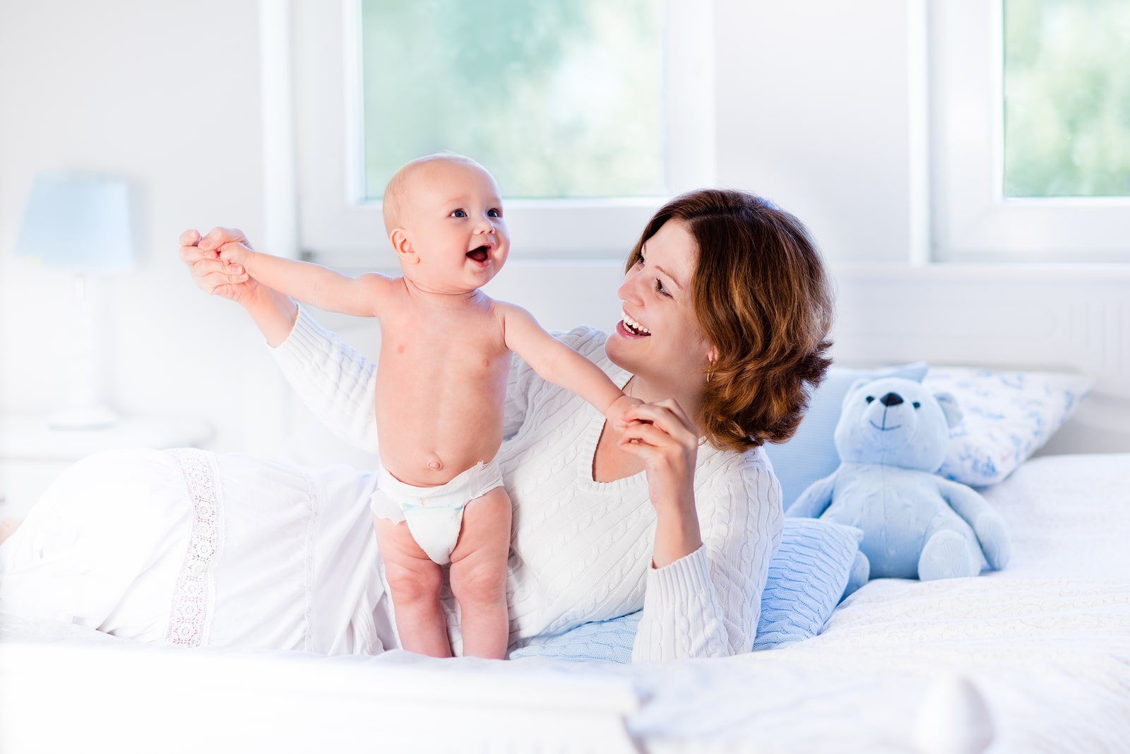 Mother and child on a white bed. Mom and baby boy in diaper playing in sunny bedroom. Parent and little kid relaxing at home. Family having fun together. Bedding and textile for infant nursery.