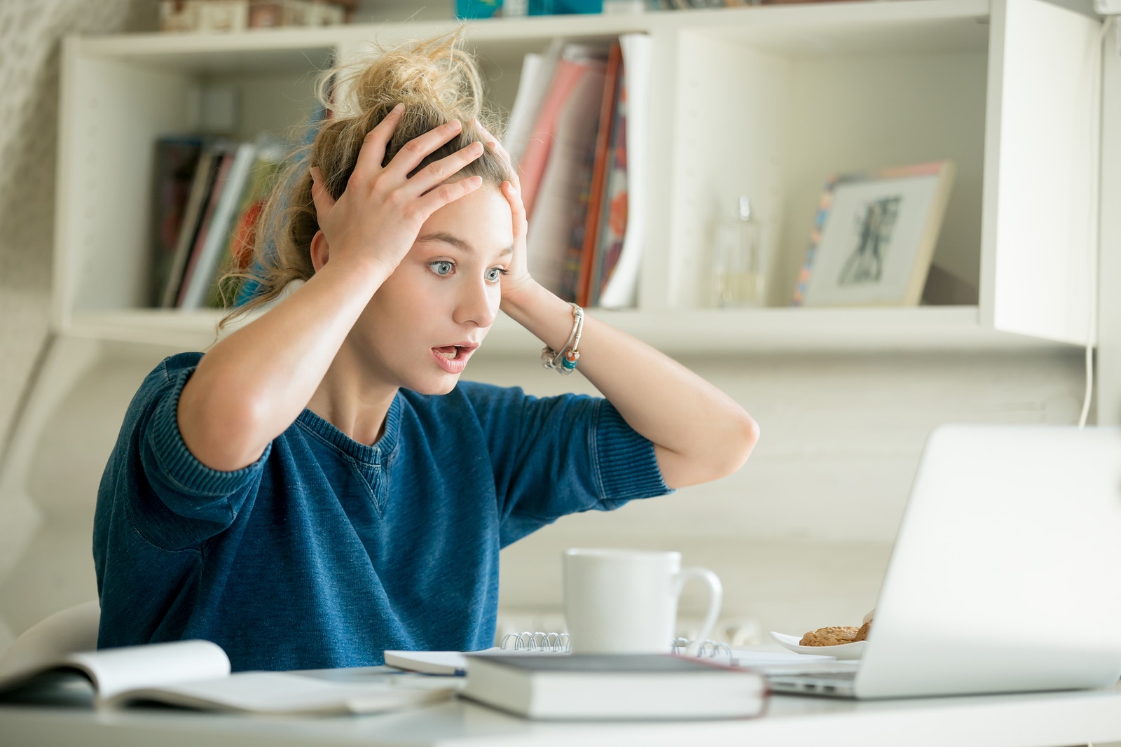 Portrait of an attractive woman at the table with cup and laptop, book, notebook on it, grabbing her head. Bookshelf at the background, concept photo