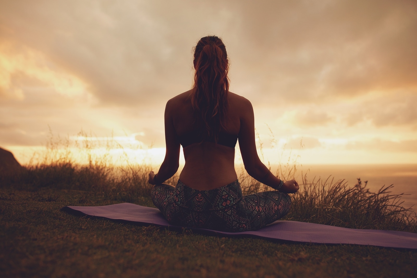Silhouette rear view of young woman doing yoga meditation outdoors. Fitness female model sitting on exercise mat in lotus yoga pose during sunset.