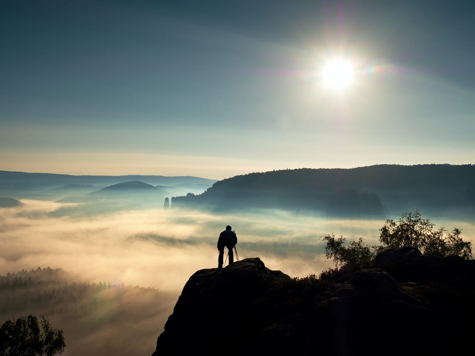 Nature photographer with tripod and camera on cliff and thinking. Dreamy fogy landscape orange misty sunrise in a beautiful mountains. Vivid and strong vignetting effect.