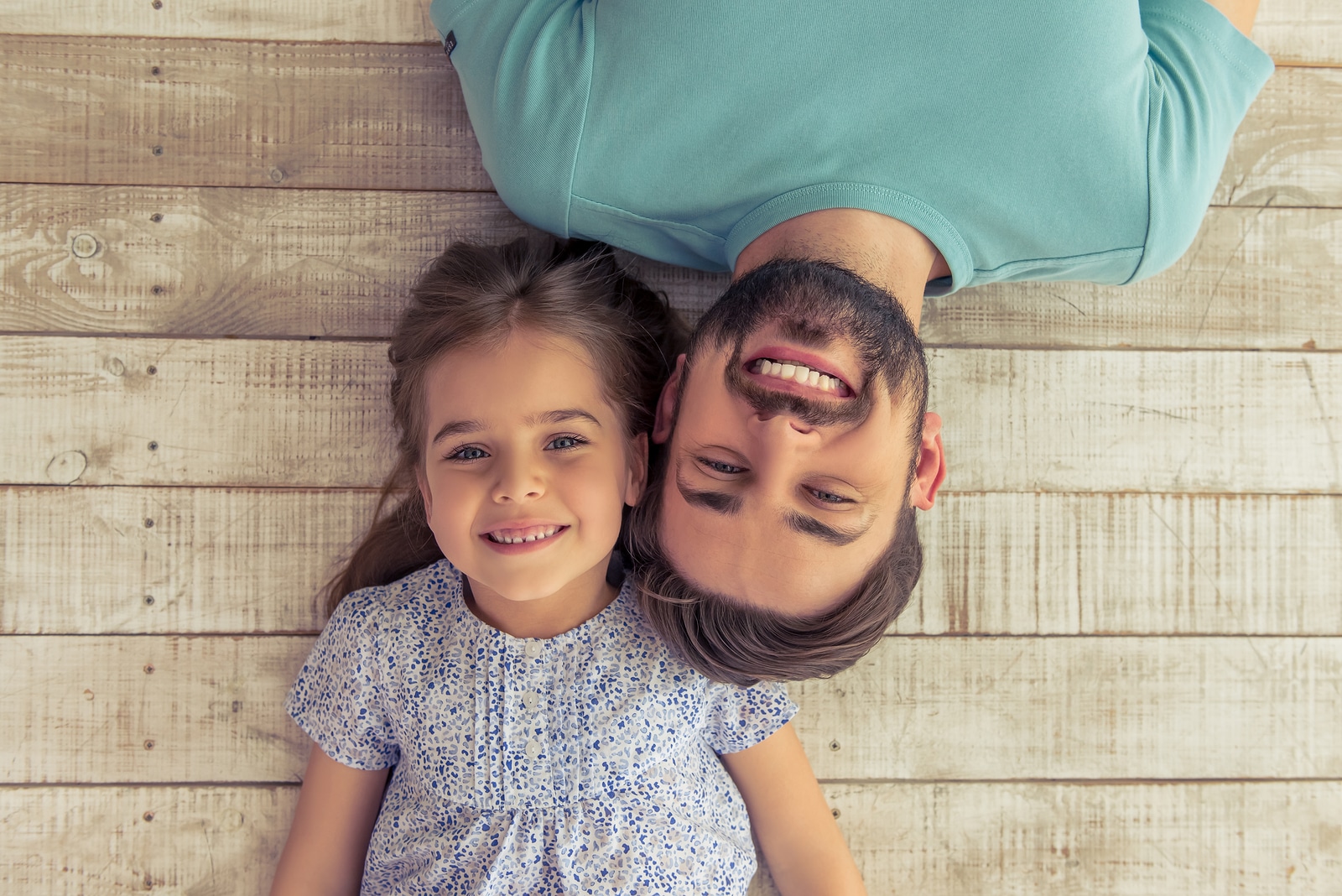 Top view of handsome young father and his cute little daughter looking at camera and smiling lying on wooden floor