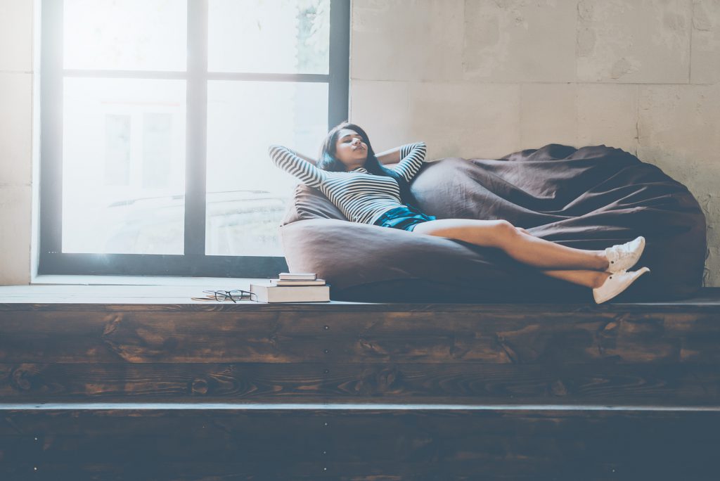 Time to relax. Beautiful young mixed race woman holding hands behind head and keeping eyes closed while lying on the comfortable couch and near the large window