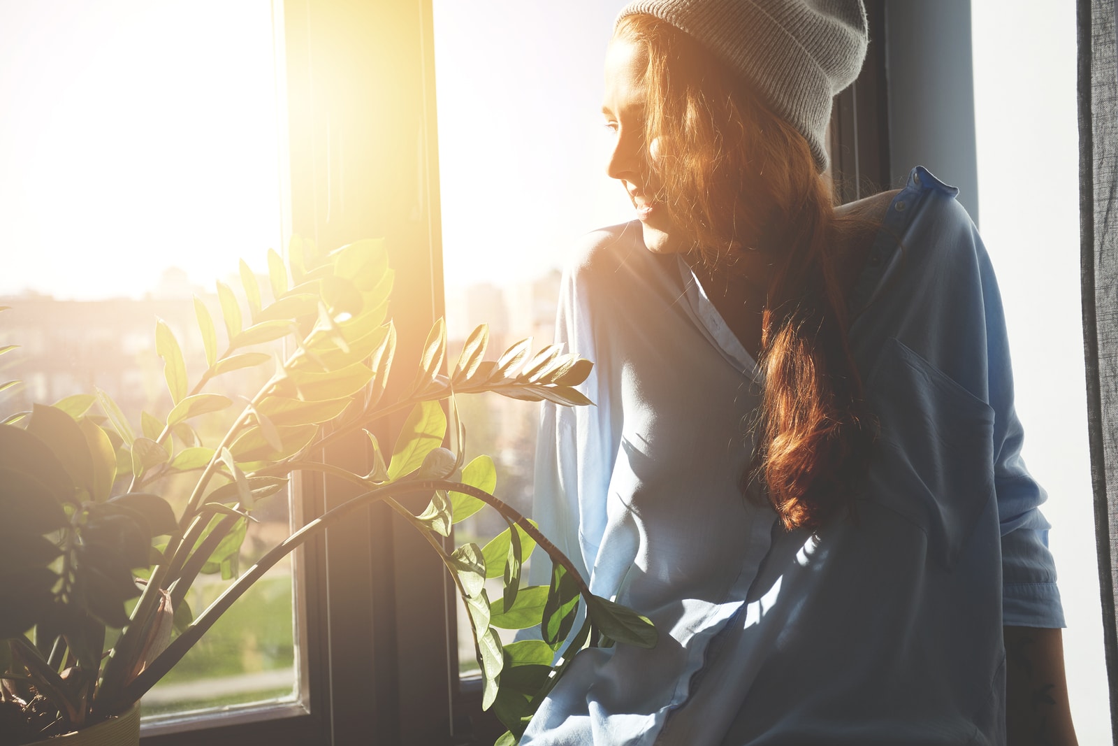 Portrait of happy Caucasian female student spending summer holidays in a big city. Hipster teenager with red hair in gray cap and shirt looking and smiling through the window. Flare sun film effect