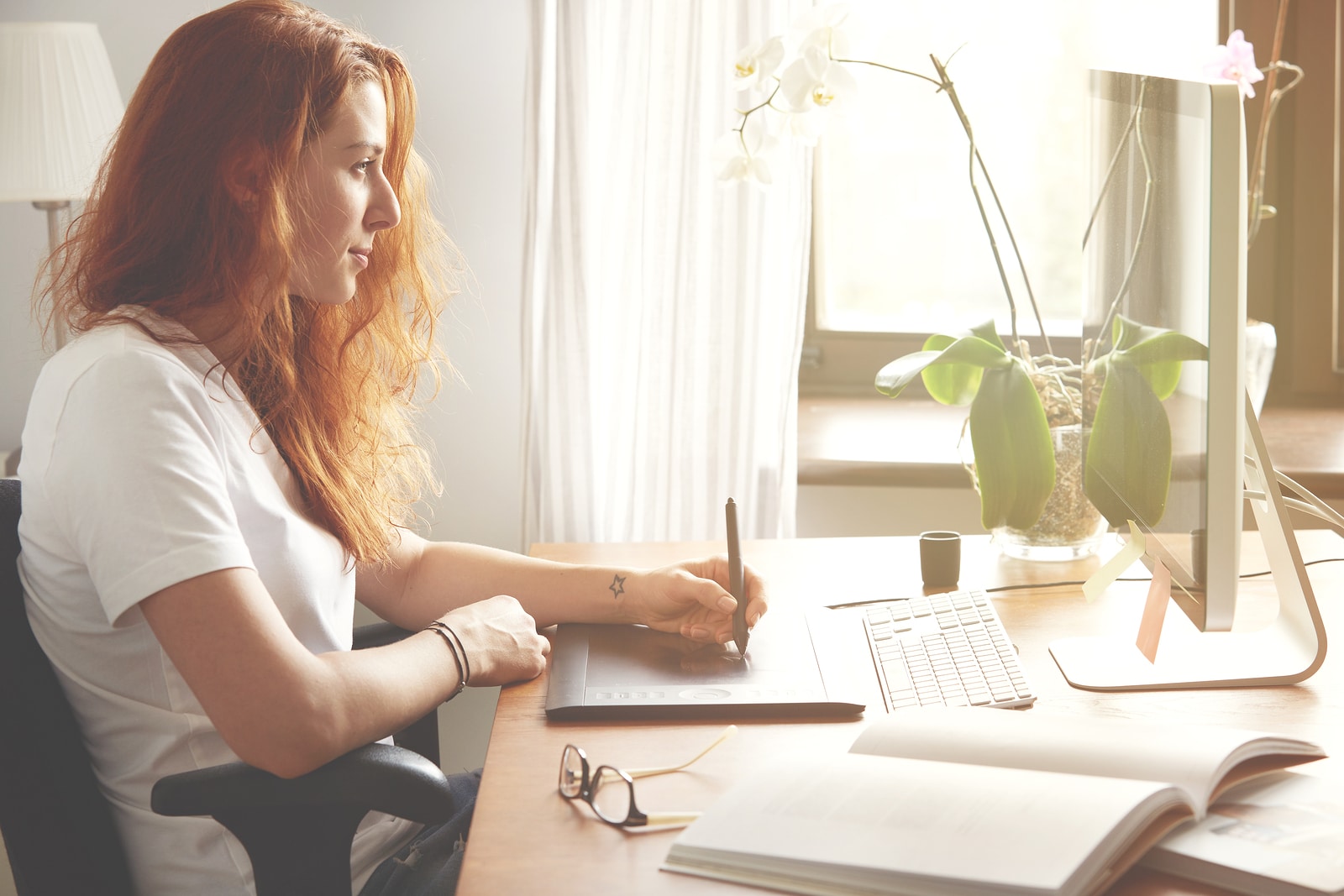 Portrait of young female freelancer working at home using digital tablet. Redhead woman designer in white T-shirt hand drawing on graphic tablet while sitting in front of computer screen. Flare sun