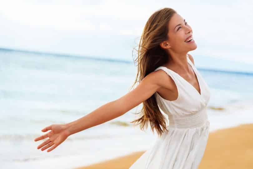 Mulher de vestido na praia, sorrindo com os braços abertos.