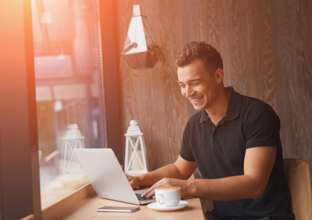 Successful entrepreneur smiling in satisfaction as he checks information on his laptop computer while working inside a cafe with sun flare