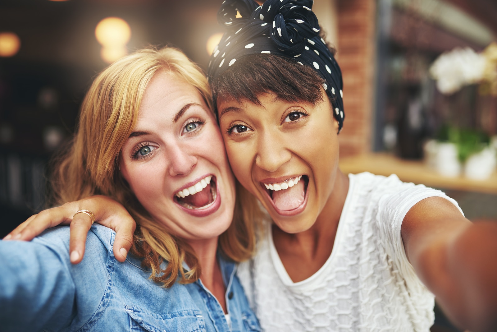 Exuberant happy multi ethnic girl friends enjoying a hearty laugh as they face the camera arm in arm inside a coffee shop head and shoulders