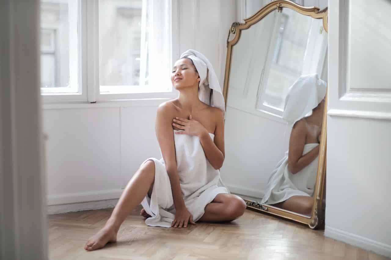 Mulher enrolada em uma toalha de banho, sentada no chão, sorrindo em frente ao espelho.