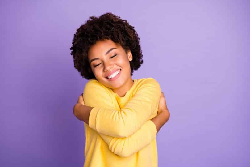 Foto de uma jovem se abraçando e sorrindo em um fundo roxo.