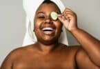 Imagem de uma mulher negra com uma toalha branca no cabelo. Ela está sorrindo, segurando uma rodela de pepino sobre um dos olhos, fazendo skincare na pele.