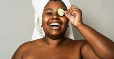 Imagem de uma mulher negra com uma toalha branca no cabelo. Ela está sorrindo, segurando uma rodela de pepino sobre um dos olhos, fazendo skincare na pele.