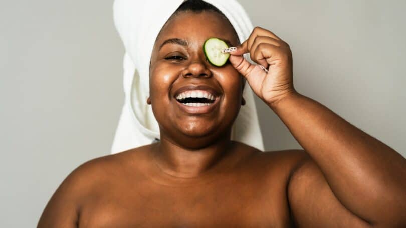 Imagem de uma mulher negra com uma toalha branca no cabelo. Ela está sorrindo, segurando uma rodela de pepino sobre um dos olhos, fazendo skincare na pele.