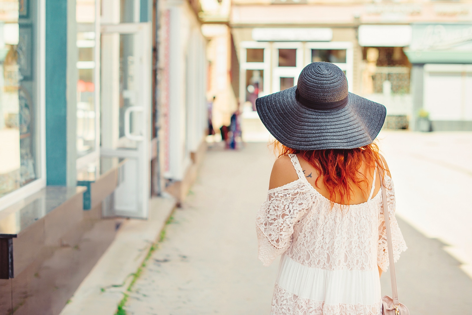 Rear view of a girl with red hair in a black hat walking around the city. Sunny summer day. City style.Girl wearing stylish summer dress of lace.