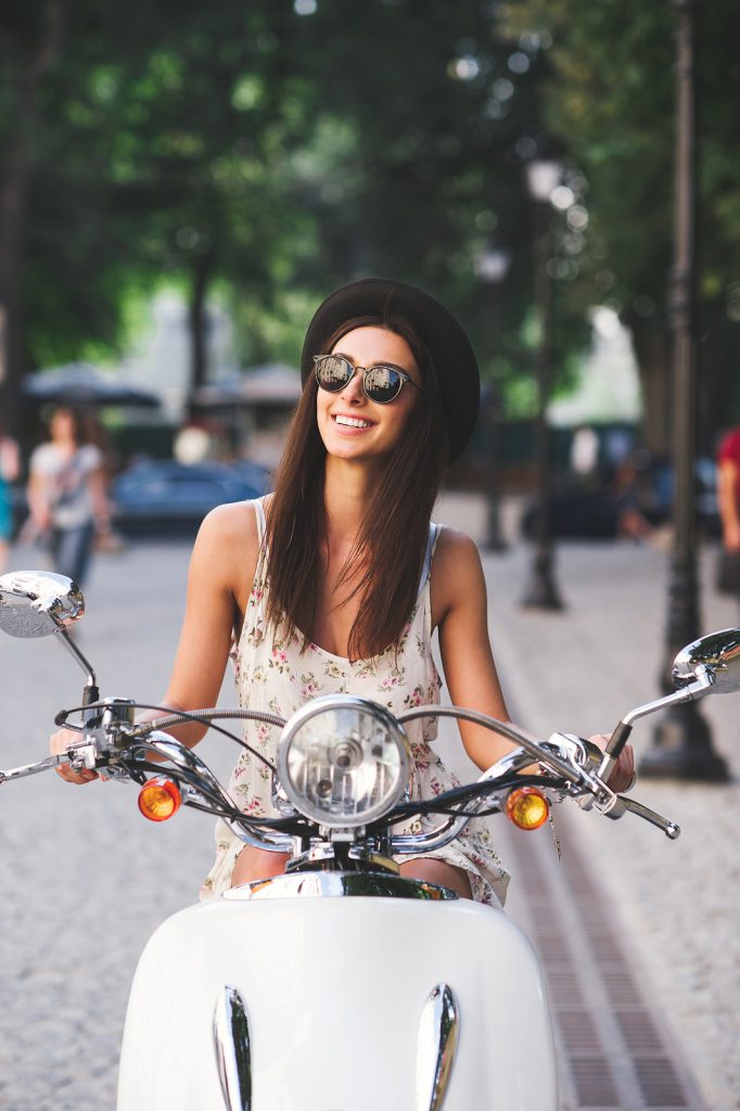 Vertical portrait of young cheerful girl driving scooter in in european city. Stylish beautiful woman in summer clothes on motorbike