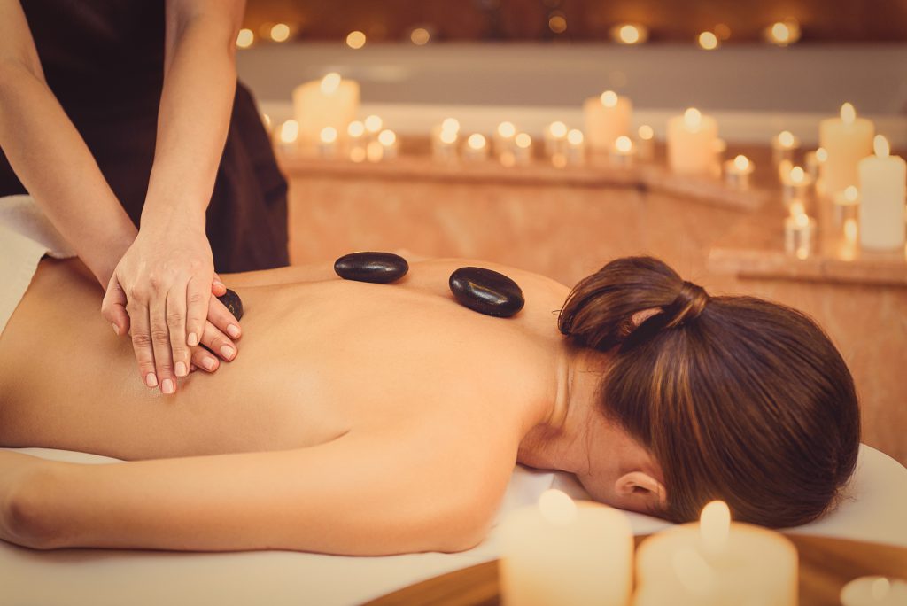 Calm young woman is getting massage at spa. Masseuse is standing and pampering her back by stone. Candles around create relaxing atmosphere